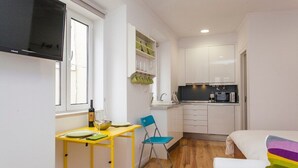 Cocina compacta con armarios blancos, microondas y un lavamanos. Hay un pequeño comedor con una mesa amarilla y una silla azul.