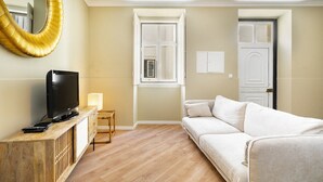 Una sala de estar con televisor sobre una consola de madera, un sofá blanco, un espejo grande y una ventana con vistas al exterior.