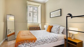 Un dormitorio con una cama, una mesita de noche con lámpara, un espejo, una ventana y un cuadro enmarcado en la pared.