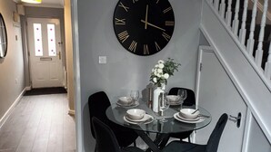 Comedor con una mesa redonda dispuesta para cuatro, un reloj de pared grande y una escalera con balaustres blancos.