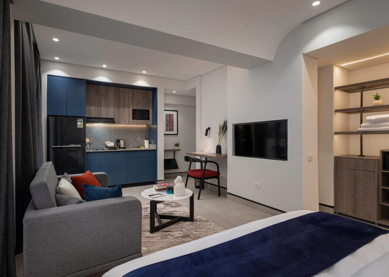 Une chambre d’hôtel moderne avec un canapé gris, une petite table ronde, un lit recouvert d’une couverture bleue, un téléviseur fixé au mur et un coin cuisine.