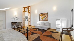 A modern hotel room with a bed, a desk with a chair, a sofa, a patterned rug, and artwork on the wall.