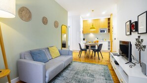 Un salon moderne avec un canapé gris, une table à manger avec des chaises, un téléviseur et une cuisine en arrière-plan.