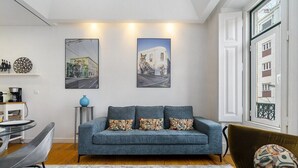 Sala de estar con sofá, una mesa de centro y un comedor con mesa y sillas. Hay dos cuadros en la pared y una ventana con vistas a edificios.
