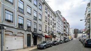Una calle empedrada flanqueada por edificios de varios pisos, coches aparcados y un escaparate con toldo rojo.