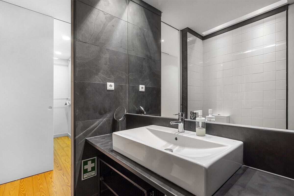 A modern bathroom with a large mirror, a white sink, and dark tiled walls.