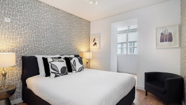 A modern bedroom with a stone-patterned wall, a large bed with black and white bedding, a black armchair, and a window with a view.