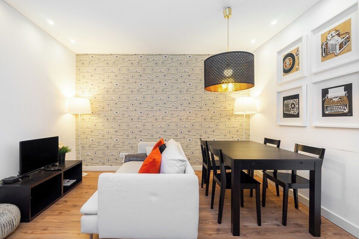 Ein modernes Wohnzimmer mit einer Couch, einem Esstisch und Stühlen. Ein Fernseher steht auf einem Schrank, es gibt eine Lampe und eine Wand mit Muster.