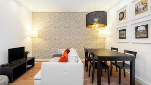 A modern living room with a sofa, a dining table, and chairs. There is a TV on a stand, a lamp, and a patterned wall.
