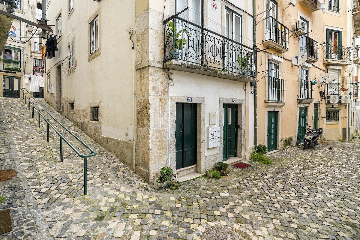 Une ruelle étroite pavée, bordée de bâtiments de chaque côté, avec des balcons ornés de rampes en métal et de petites plantes.