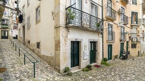 A narrow cobblestone street with buildings on either side, featuring balconies with metal railings and small plants.