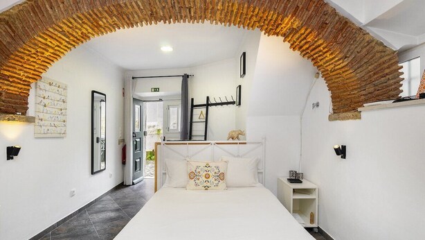 A bedroom with a white bed, a decorative pillow, a headboard, a small bedside table, and a wall-mounted shelf.
