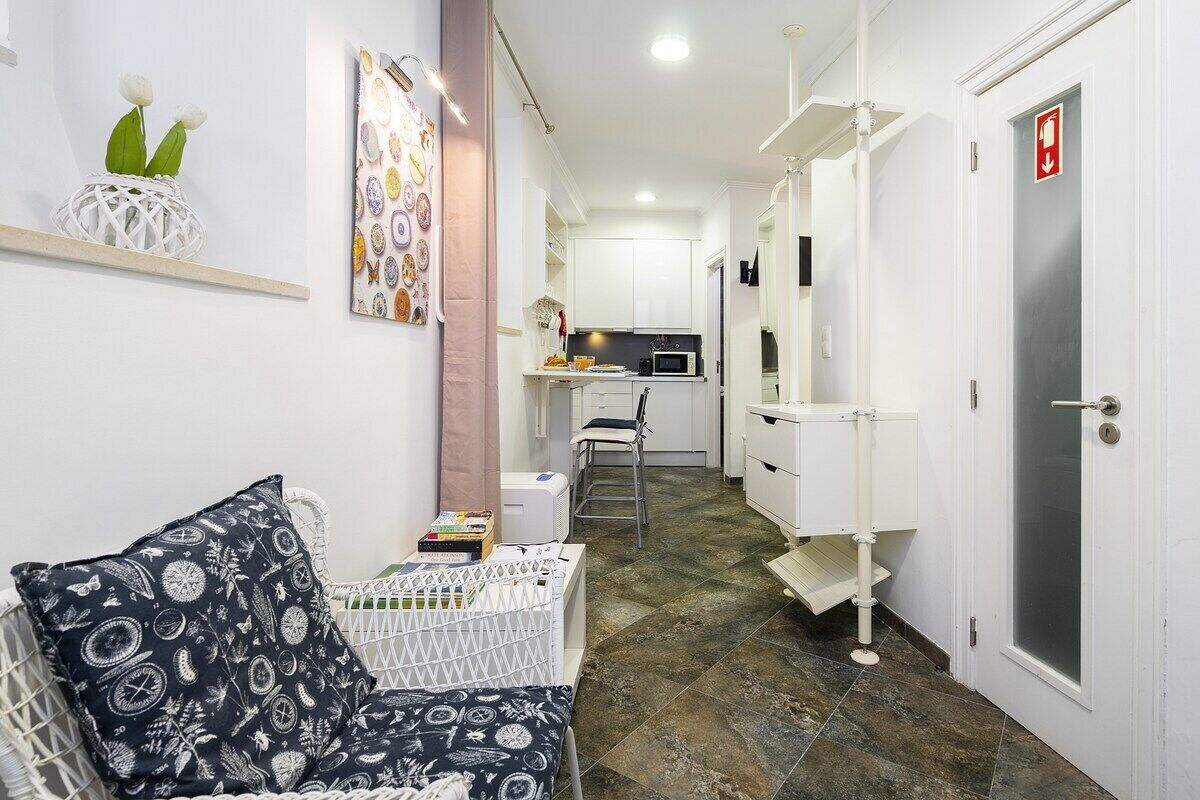 A narrow hallway with a white door on the right, a white chair, and a small kitchenette area with a microwave and sink.