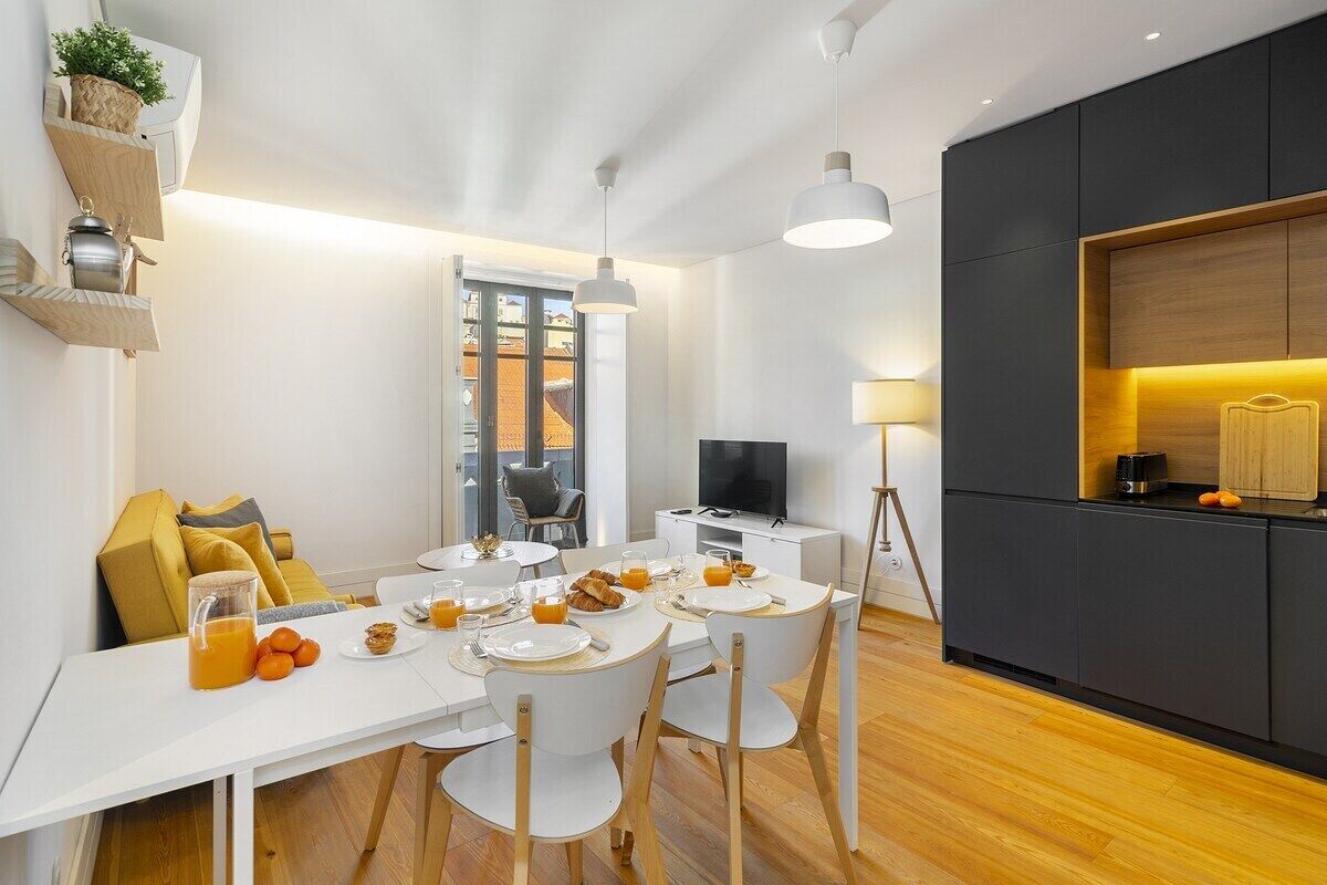 Un comedor moderno con una mesa blanca dispuesta para el desayuno, una cocina con armarios oscuros y vistas a la ciudad a través de un ventanal.