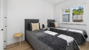 A bedroom with two beds, a small side table, a lamp, and a window with a view of the outside. - Suburban Serenity: Family Home with Pool (Algés)