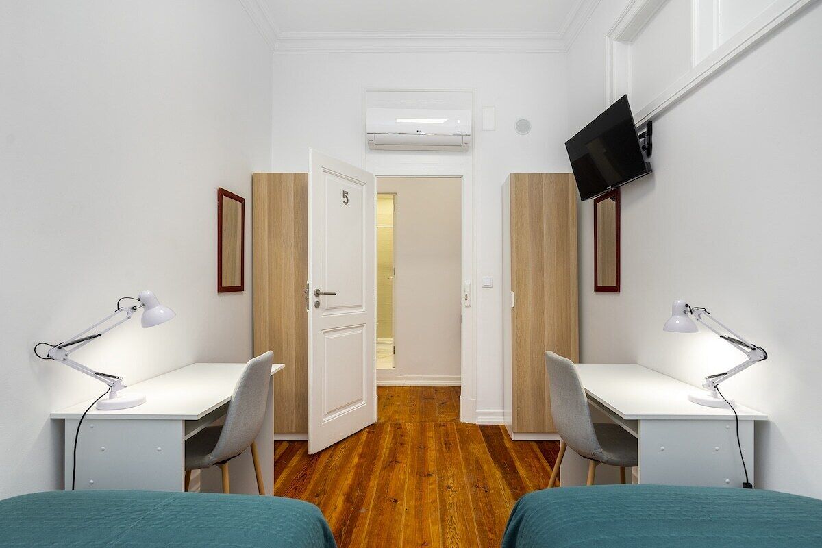 Una habitación con dos escritorios blancos, cada uno con una lámpara de escritorio blanca, una cama con ropa de cama color turquesa y un televisor montado en la pared.