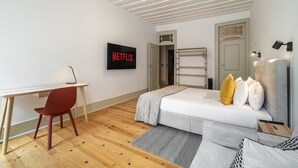 Une chambre moderne avec un plancher en bois, un lit recouvert d’une couette grise, un bureau avec une chaise, un téléviseur fixé au mur et une échelle appuyée contre le mur.
