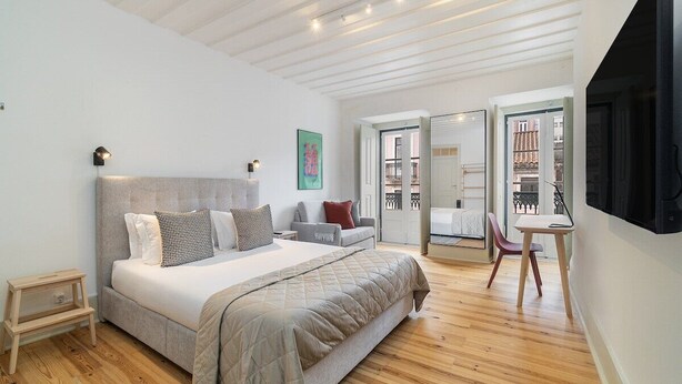 A bedroom with a large bed, a small table, and a chair. There is a door leading to a balcony. - Lisbon's Heartbeat: Your Premier Airbnb 4Explorers (Lisboa)
