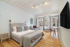 Une chambre avec un grand lit, une petite table et une chaise. Une porte donne sur un balcon.