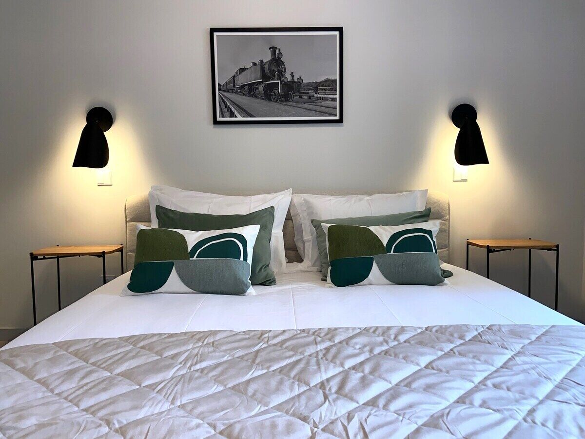 Uma cama bem arrumada com um edredom branco acolchoado, duas mesas de cabeceira com abajures pretos e uma fotografia a preto e branco emoldurada na parede.