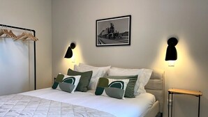 A neatly made bed with a quilted white duvet, a headboard with a light-colored fabric, and a framed black and white photograph above it. There are two wall-mounted black light fixtures on either side of the bed.