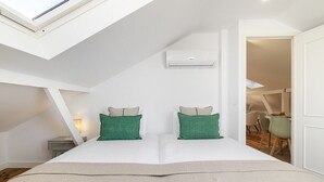 Una camera da letto con un lucernario, un letto con cuscini verde e beige, un condizionatore d'aria bianco e vista su una sala da pranzo.
