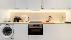 Une cuisine compacte équipée d’un lave-linge, d’une machine à café, d’une bouilloire et d’une planche à découper avec une tomate.