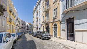 Une rue pavée bordée d’immeubles à plusieurs étages, de voitures garées et d’un ciel d’un bleu éclatant.