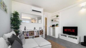 Ett modernt vardagsrum med en vit soffa, en platt-TV som visar ”NETFLIX” och en bokhylla med diverse föremål.