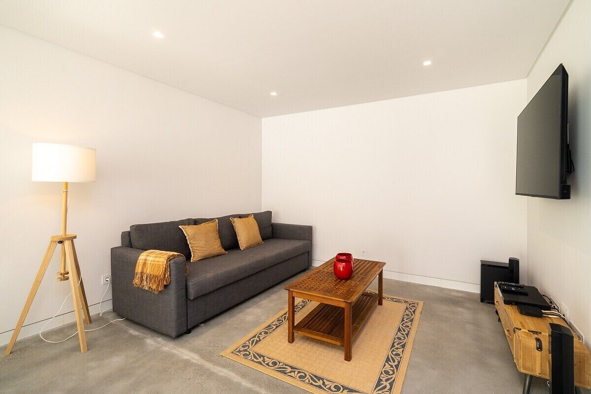 Uma sala de estar moderna com um sofá cinzento, uma mesa de centro de madeira, um abajur e uma televisão de tela plana fixada na parede.