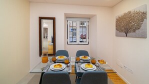 Zona pranzo con tavolo in vetro apparecchiato per quattro, una finestra con persiane e un quadro appeso al muro.
