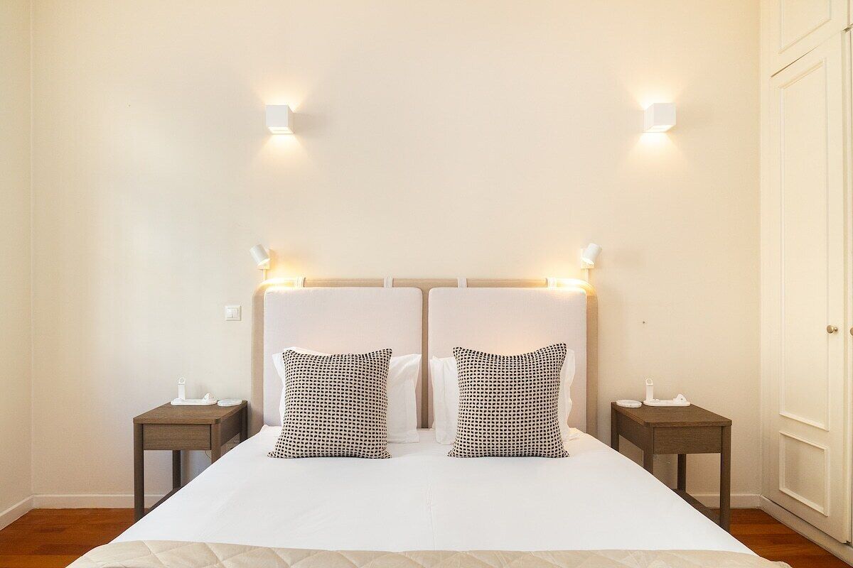 Uma cama de casal com edredom branco, duas almofadas estampadas e duas mesas de cabeceira com abajures.