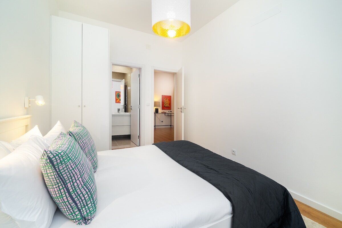 Un dormitorio con cama, paredes blancas, una puerta que da a un baño y una lámpara en el techo.