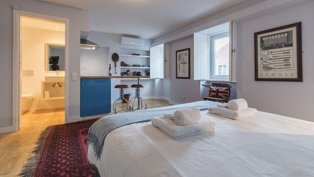 A modern bedroom with a large bed, a sitting area with stools, a kitchen area, and a bathroom. - Tennis Room - Clean & Safe (Lisboa)