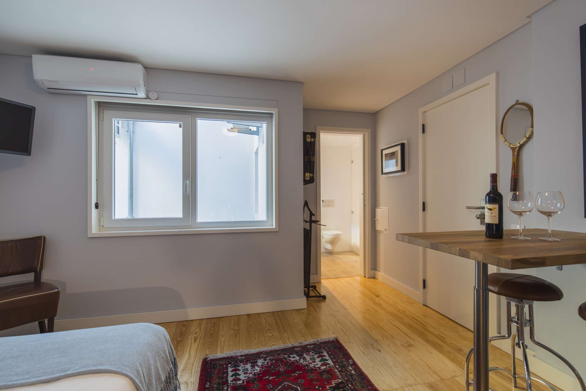 Una stanza moderna con un'ampia finestra, un tavolo in legno con una bottiglia di vino e calici, uno sgabello, una TV a parete e una porta che conduce al bagno.
