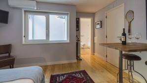 Ein modernes Zimmer mit einem großen Fenster, einem Holztisch mit Weinflasche und Gläsern, einem kleinen Hocker, einem an der Wand montierten Fernseher und einer Tür zu einem Badezimmer.