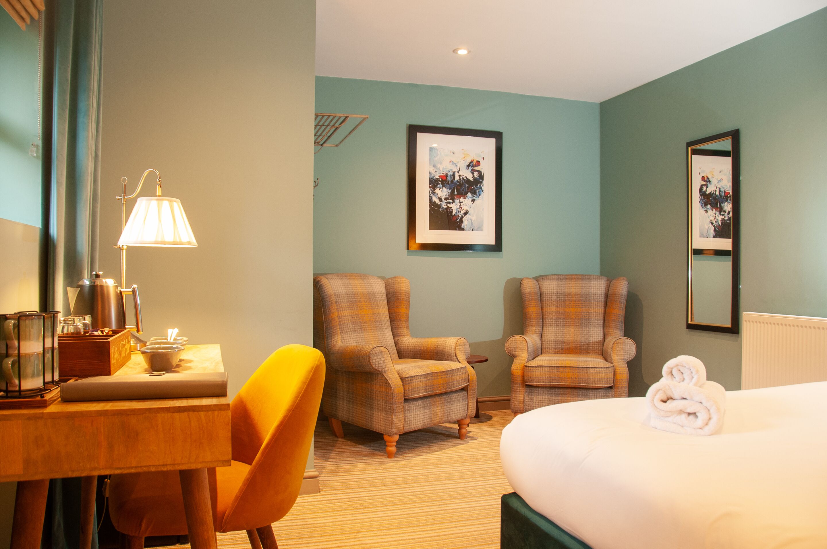 Habitación de hotel con cama, dos sillones, un escritorio con lámpara y obras de arte en las paredes.