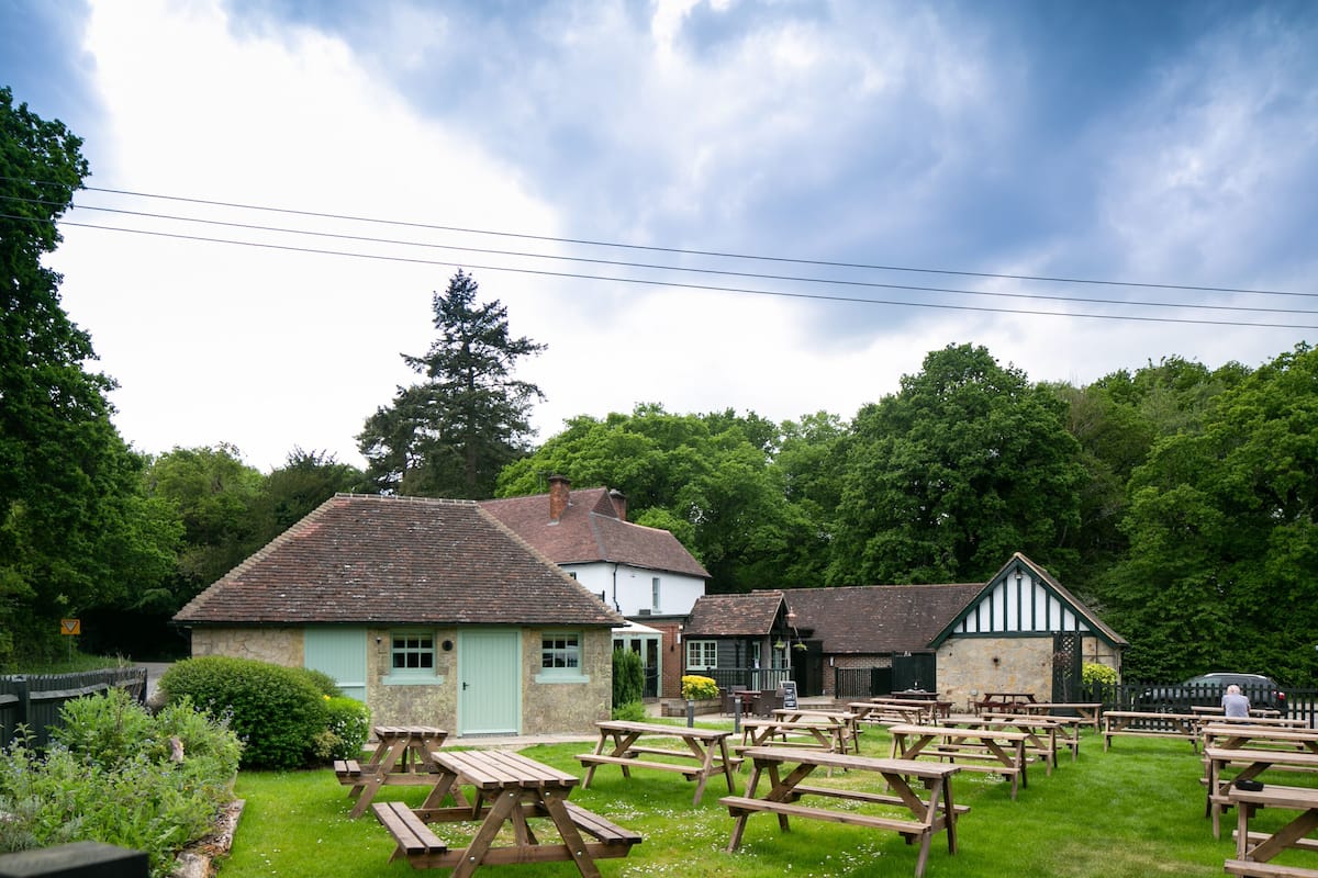 A rustic outdoor seating area with picnic tables, surrounded by traditional buildings with stone walls and pitched roofs.