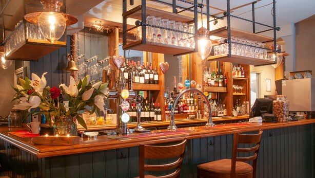 A bar area with wooden bar counter, multiple stools, shelves with bottles, and hanging glassware.