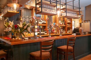 A bar area with wooden bar counter, multiple stools, shelves with bottles, and hanging glassware. - Cowdray Arms (Haywards Heath)