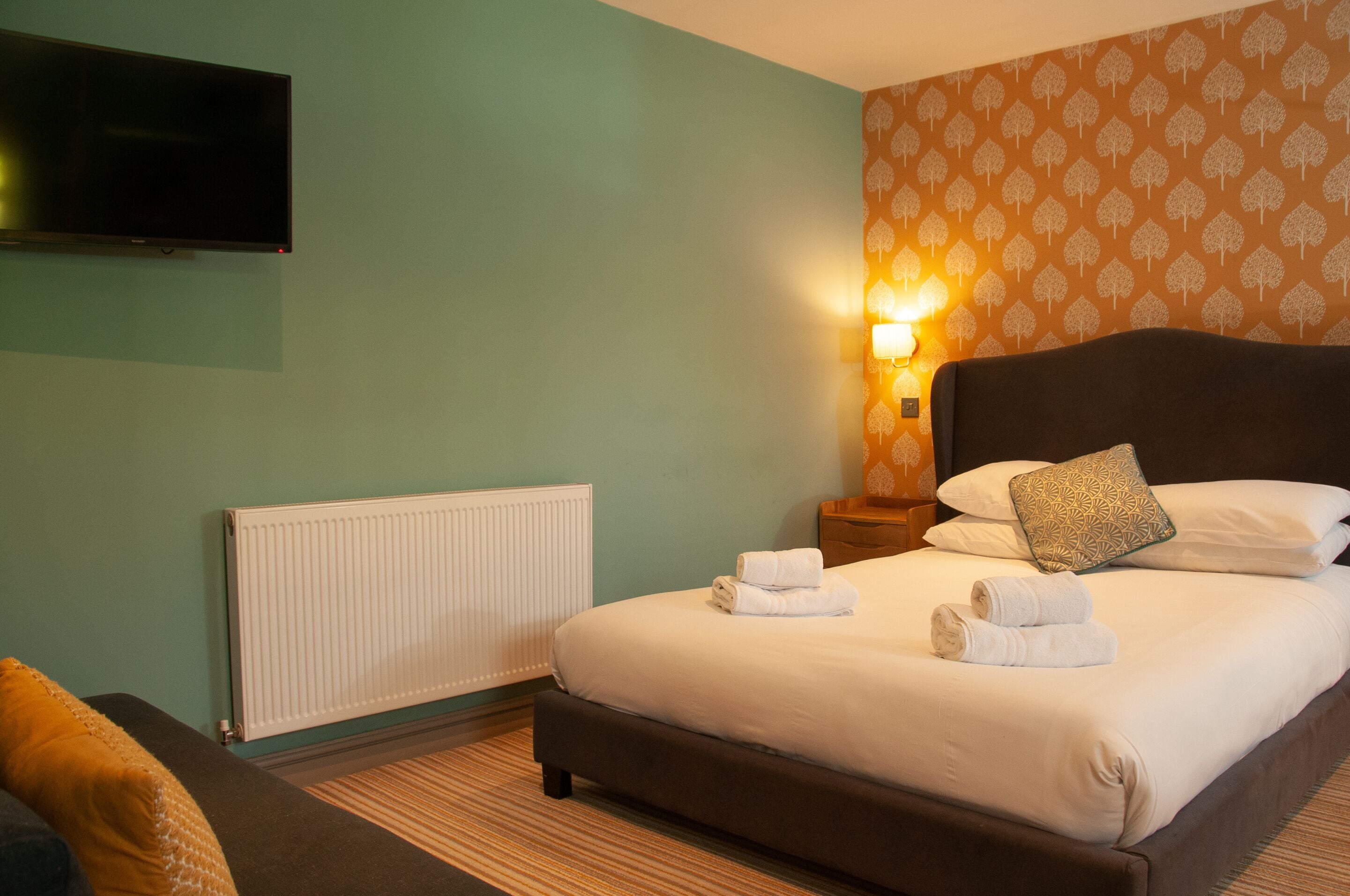 A hotel room with a bed, a TV, a wall-mounted light, and a radiator.