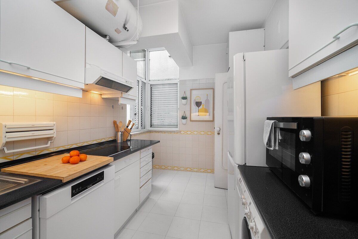 Cocina compacta con armarios blancos, encimera negra, tabla de cortar de madera con naranjas, microondas y ventana con persianas.