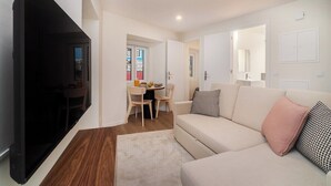 A modern living room with a sofa, a flat-screen TV, a dining area with a table and chairs, and a hallway leading to another room.