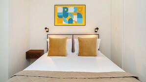 Una cama bien hecha con edredón blanco y dos almohadas doradas, una mesita de noche de madera y un cuadro abstracto colgado en la pared.