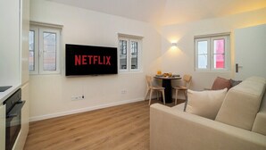 A modern living room with a flat-screen TV mounted on the wall, a sofa, a dining table with chairs, and a kitchen area with a stove. - Explore Lisbon from a Cozy Base in Alfama (Lisboa)