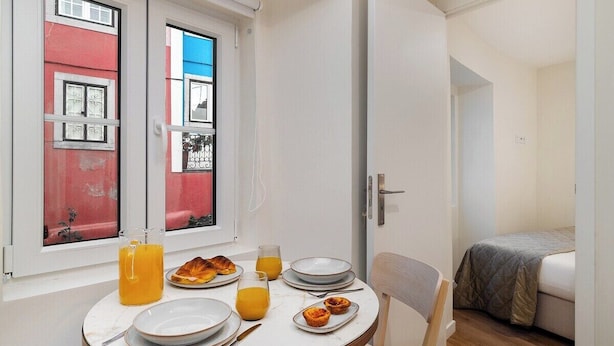 A small, well-lit room with a round dining table set for breakfast, a bed with a grey blanket, and a window with a view of a colorful building. - Explore Lisbon from a Cozy Base in Alfama (Lisboa)
