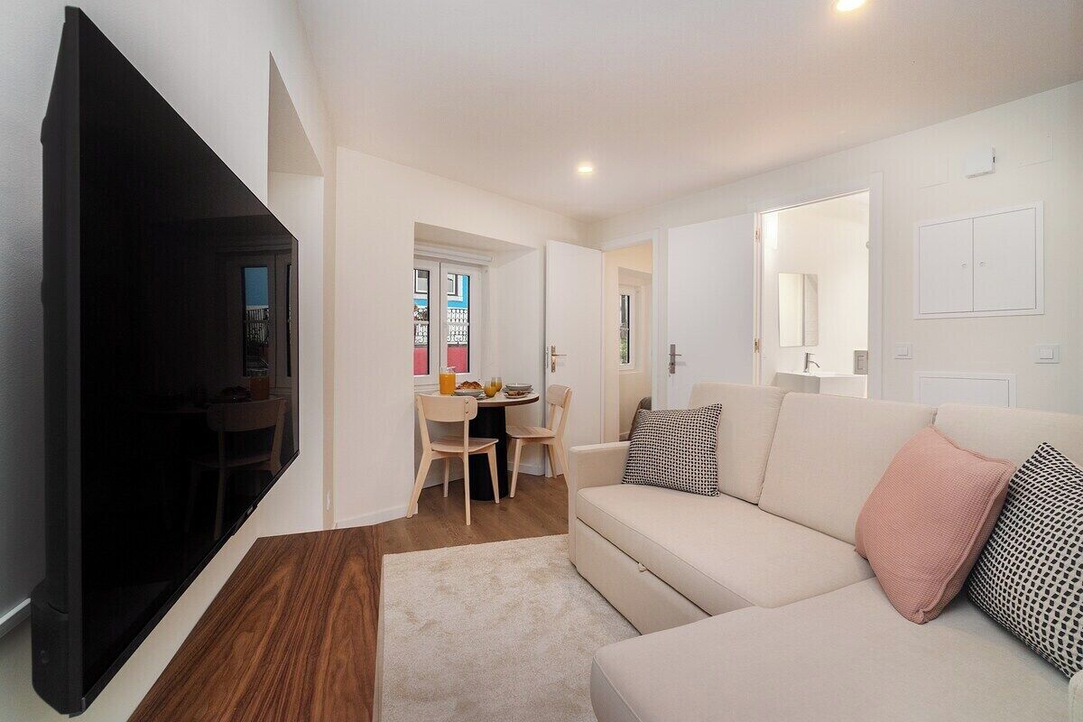 A modern living room with a sofa, a flat-screen TV, a dining area with a table and chairs, and a hallway leading to another room.