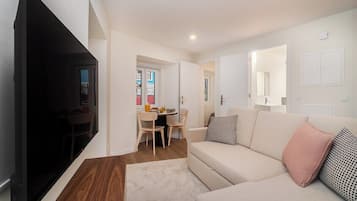 A modern living room with a sofa, a flat-screen TV, a dining area with a table and chairs, and a hallway leading to another room.
