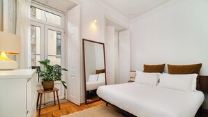 A bedroom with a large bed, a bedside table, a mirror, a potted plant, and a window with curtains.