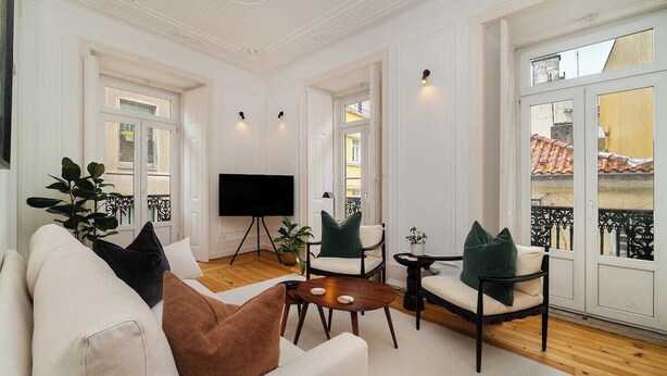 A spacious living room with a white sofa, a coffee table, and two armchairs. A flat-screen TV is mounted on the wall, and there are large windows with a view of the city.